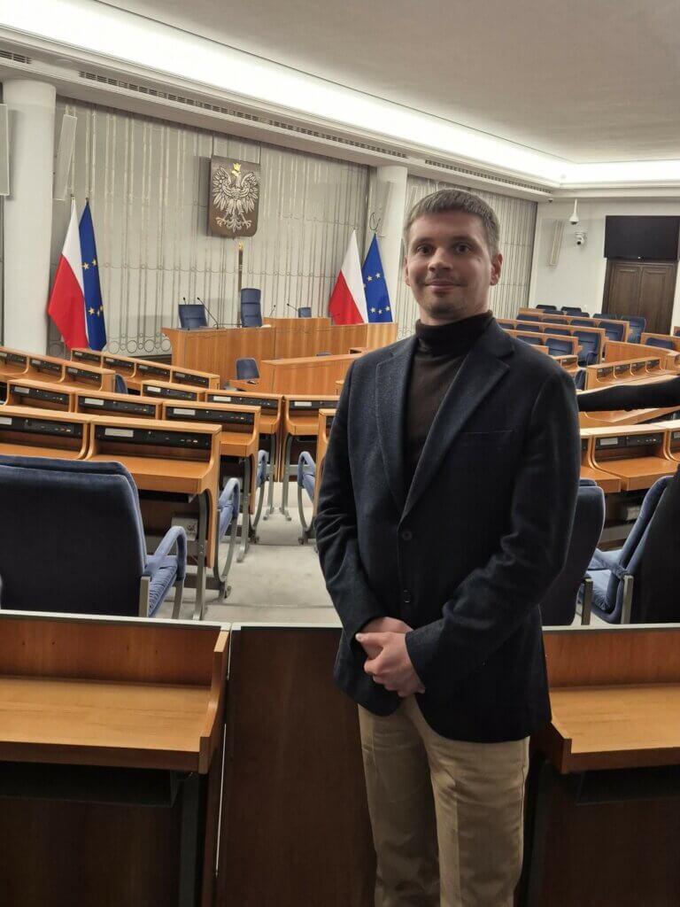 Łukasz Ptak w sali obrad Senatu RP z flagami Polski i Unii Europejskiej